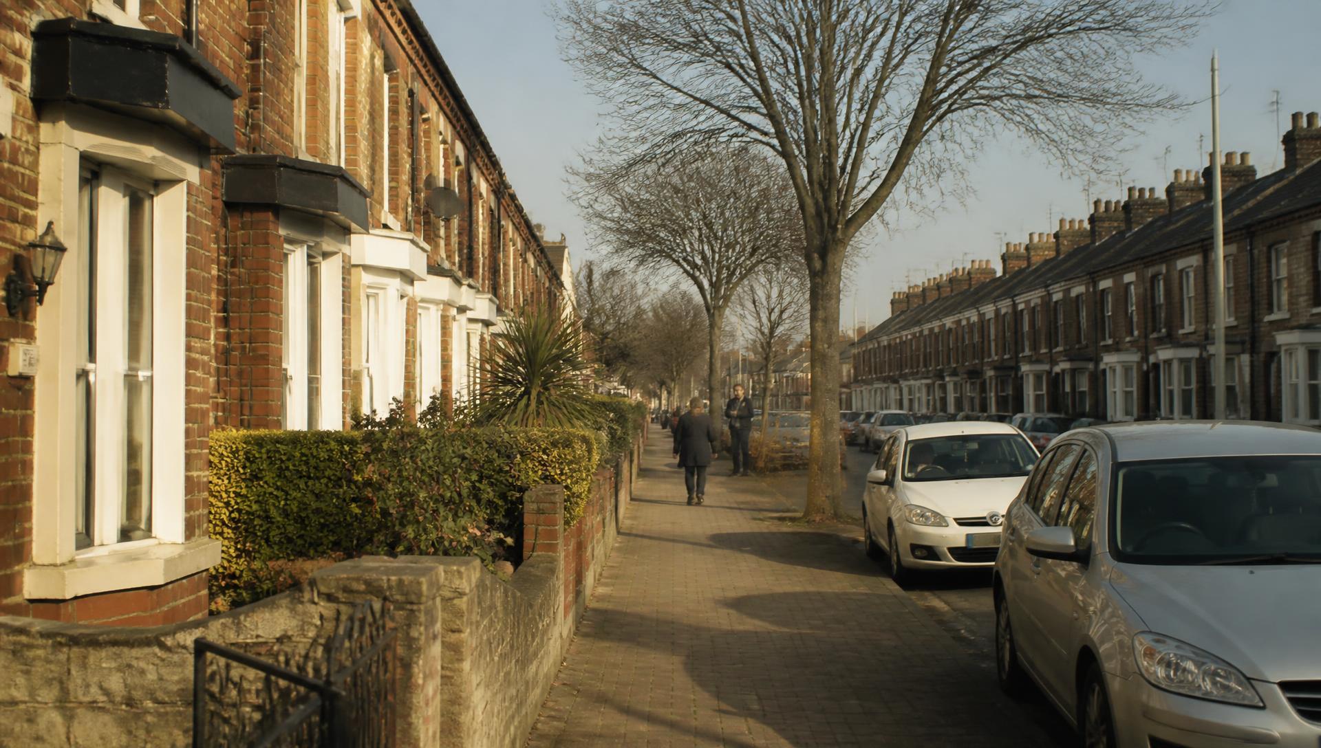 A quiet residential street of modest brick homes in morning light, with neighbours walking nearby